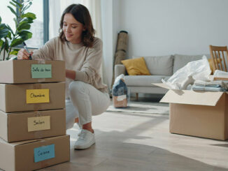 Déballage de cartons : image ou photo affichant une pile de cartons bien rangés au premier plan. Chaque carton possède une étiquette colorée visible (Cuisine, Salon, Chambre, Salle de bain). Une personne souriante tient un marqueur et termine d’étiqueter un carton.