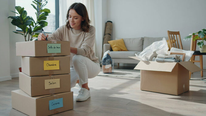 Déballage de cartons : image ou photo affichant une pile de cartons bien rangés au premier plan. Chaque carton possède une étiquette colorée visible (Cuisine, Salon, Chambre, Salle de bain). Une personne souriante tient un marqueur et termine d’étiqueter un carton.