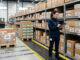 Audit stockage : image ou photo d'un entrepôt propre et bien organisé, avec des cartons d’emballage empilés sur des étagères. Au premier plan, une personne tient une tablette ou une feuille de contrôle, comme si elle faisait un audit du stock. On voit aussi quelques palettes, des zones de rangement bien délimitées.